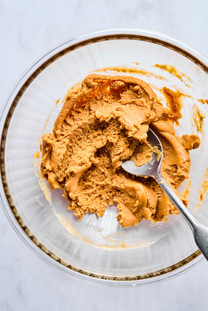 A glass bowl of dough with a spoon in it.