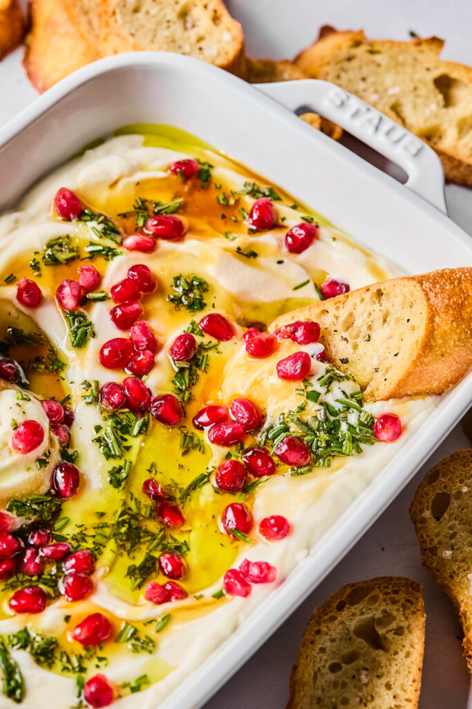 Whipped ricotta dip in a white serving dish topped with olive oil, fresh rosemary, pomegranate seeds, and hot honey. A piece of crostini is dipped into the ricotta and around the pan is more crostini.