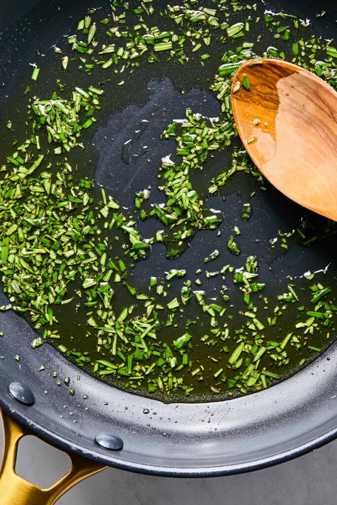 Overhead shot of a skillet with chopped rosemary being cooking in olive oil. A wooden spoon is also in the skillet.