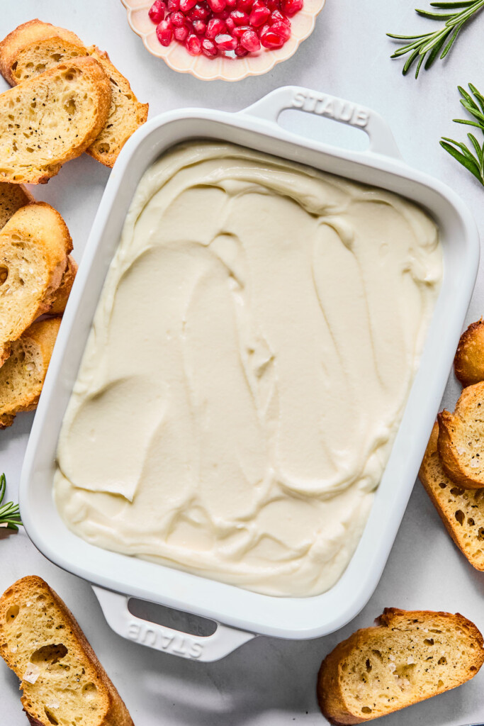 Small serving dish with whipped ricotta cheese in it. Around the dish are some crostini, a bowl of pomegranate seeds, and a couple of sprigs of fresh rosemary.
