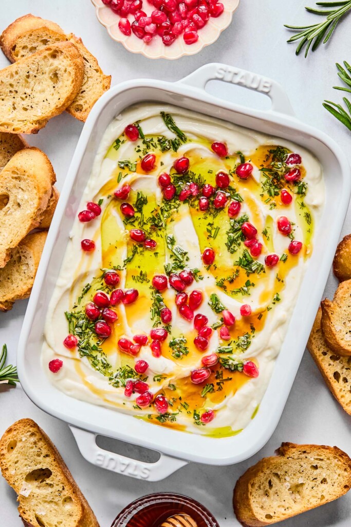 Overhead shot of whipped ricotta topped with olive oil, rosemary, pomegranates, and hot honey in a small dish. Around the dish are some crostini, a bowl of pomegranate seeds, and a couple of sprigs of fresh rosemary.