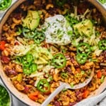 Overhead shot of chili pasta in a large Dutch oven. It is topped with sour cream, shredded cheese, sliced avocado, sliced jalapeños, sliced green onions, and cilantro. A metal spoon is also in the pot. Around the pot is a striped kitchen towel, a bowl of sliced green onions, a bowl of sliced jalapeños, a bowl of shredded cheese, and a bowl of chopped cilantro.