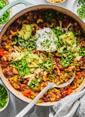 Overhead shot of chili pasta in a large Dutch oven. It is topped with sour cream, shredded cheese, sliced avocado, sliced jalapeños, sliced green onions, and cilantro. A metal spoon is also in the pot. Around the pot is a striped kitchen towel, a bowl of sliced green onions, a bowl of sliced jalapeños, a bowl of shredded cheese, and a bowl of chopped cilantro.