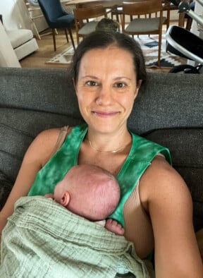 Selfie of a mom sitting on the couch with her baby napping on her. There is a blanket on the baby.
