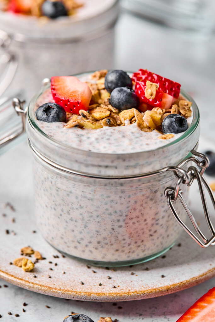 A jar of protein chia pudding topped with strawberries, blueberries, and granola. The jar is sitting on a plate and there is another jar filled with chia pudding behind the jar.