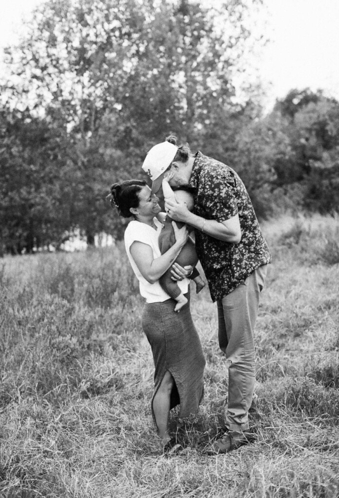 A mother and father standing in a field holding their baby together. The dad is kissing the top of the baby's head and the mom is smiling at them.