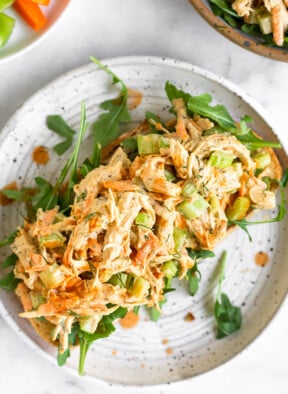 Healthy chicken salad with celery, carrots, and buffalo sauce on top of some arugula on a piece of bread on a plate. It is drizzled with hot sauce. Around the plate is a plate of carrots and celery and a bowl of more chicken salad.