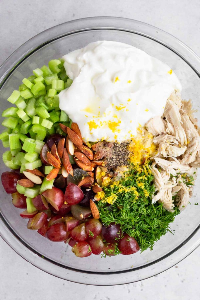 A large glass bowl of greek yogurt, shredded chicken, fresh dill, chopped grapes, chopped almonds, diced celery, lemon zest, and salt and pepper before it is mixed together.