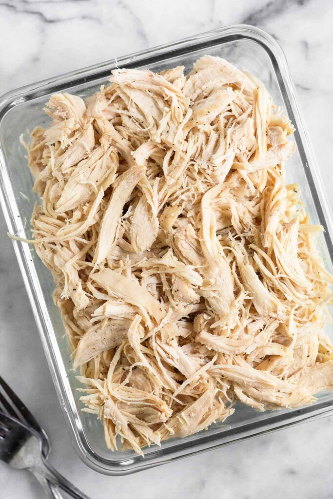 Glass rectangular container filled with shredded chicken seasoned with salt and pepper. It is on a marble counter with two forks next to the dish.