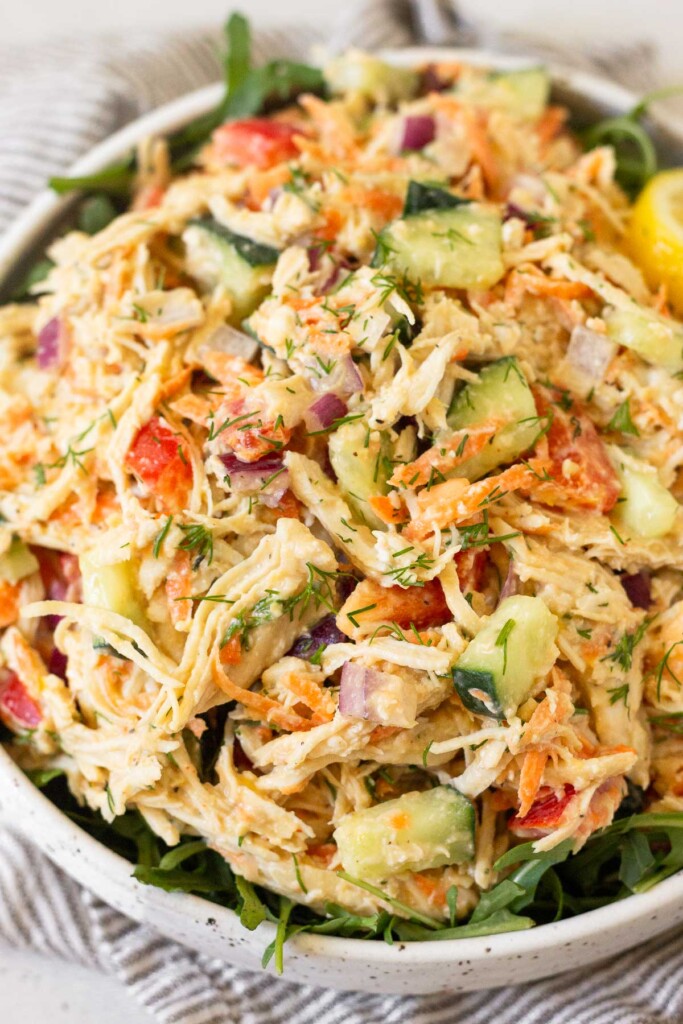 Hummus chicken salad with shredded carrots, diced bell pepper, diced cucumber, and fresh dill in a hummus dressing overtop greens in a white bowl.