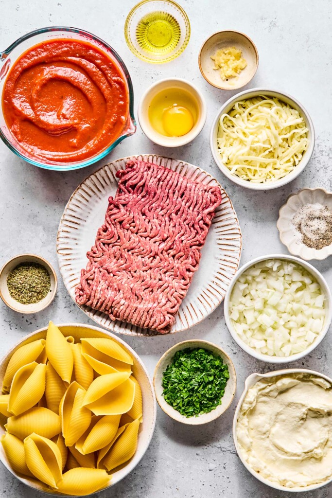 A small bowl of olive oil, a small bowl of minced garlic, a small dish with a cracked raw egg in it, a bowl of shredded mozzarella cheese, a dish with salt and pepper in it, a bowl of diced onions, a bowl of ricotta cheese, a bowl of chopped parsley, a bowl of uncooked jumbo pasta shells, a small bowl with Italian seasoning, a plate with a pound of raw ground beef, and a large measuring cup of pasta sauce on a grey counter.