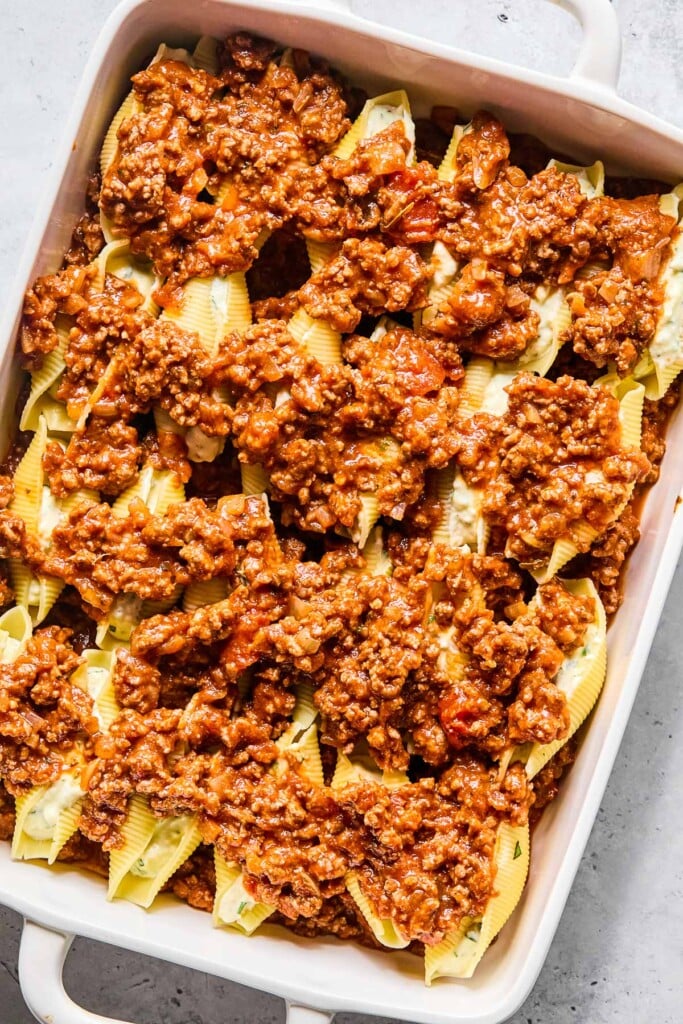 A large baking dish with meat sauce spread across the bottom with pasta stuffed shells on top of it. On top of the shells is more meat sauce.
