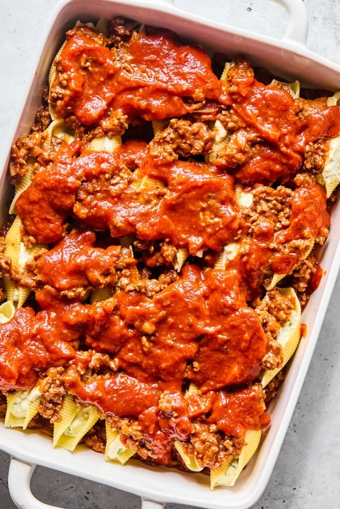 A large baking dish with meat sauce spread across the bottom with pasta stuffed shells on top of it. On top of the shells is more meat sauce and pasta sauce.