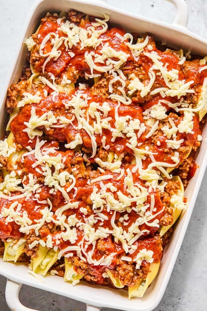 A large baking dish with meat sauce spread across the bottom with pasta stuffed shells on top of it. On top of the shells is more meat sauce, pasta sauce, and shredded mozzarella cheese.