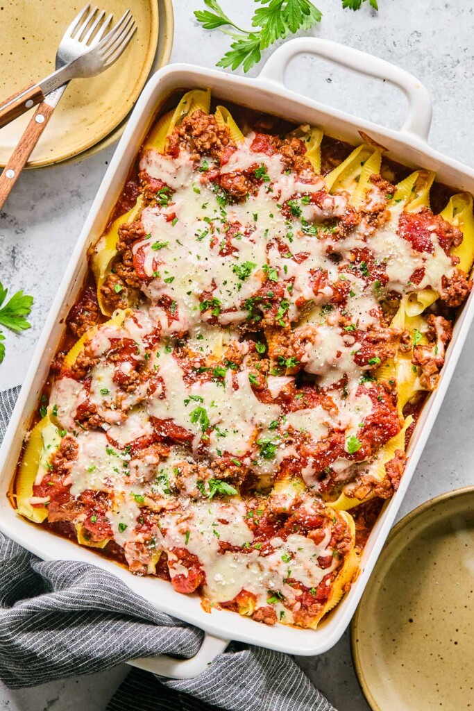 Stuffed shells with ground beef sauce in a pan. They are topped with melted cheese, fresh parsley, and grated parmesan cheese. Around the pan are bowls, a kitchen towel, two forks, and fresh parsley.