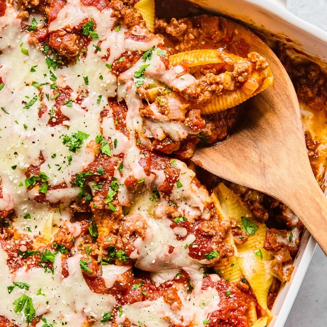 Overhead shot of a pan filled with stuffed shells with meat sauce topped with melted cheese, parsley, and parmesan cheese. There is a wooden spoon in the pan with a stuffed shell on it.
