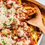 Overhead shot of a pan filled with stuffed shells with meat sauce topped with melted cheese, parsley, and parmesan cheese. There is a wooden spoon in the pan with a stuffed shell on it.