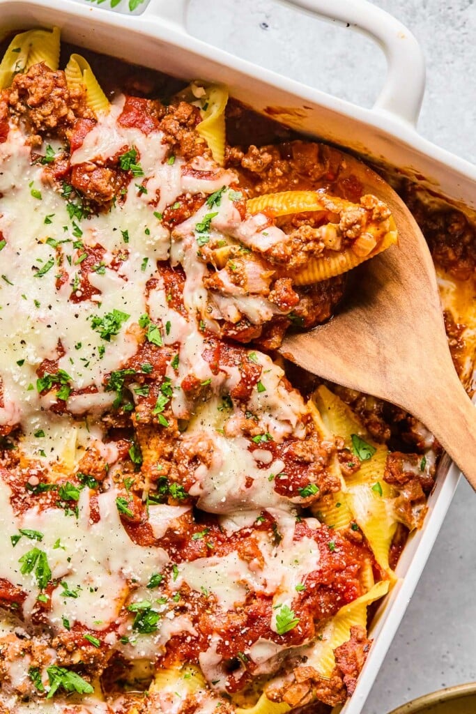 Overhead shot of a pan filled with stuffed shells with meat sauce topped with melted cheese, parsley, and parmesan cheese. There is a wooden spoon in the pan with a stuffed shell on it.