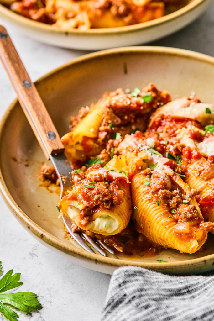 A bowl of stuffed shells with meat and ricotta topped with fresh parsley and parmesan cheese. A fork is also in the bowl. Around the bowl is a kitchen towel, more fresh parsley, and a bowl of more stuffed shells.