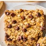 A piece of banana baked oatmeal with chocolate chips on a white plate. A little piece is taken off the corner and sitting on a fork on the plate. Around the plate are some chocolate chips and some bananas.