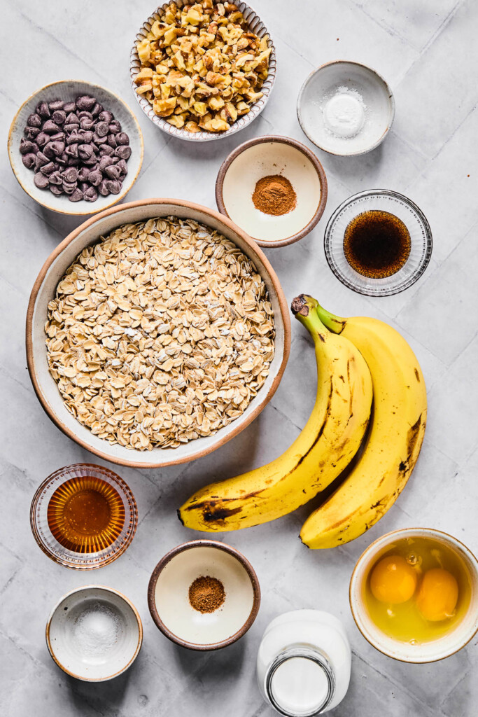 A bowl of walnuts, a small bowl of baking powder, a small bowl of cinnamon, a small bowl of vanilla bean paste, two ripe bananas, a bowl with two cracked eggs in it, a jar of milk, a small bowl of nutmeg, a small bowl of salt, a bowl of honey, a large bowl of rolled oats, and a bowl of chocolate chips on a white counter.