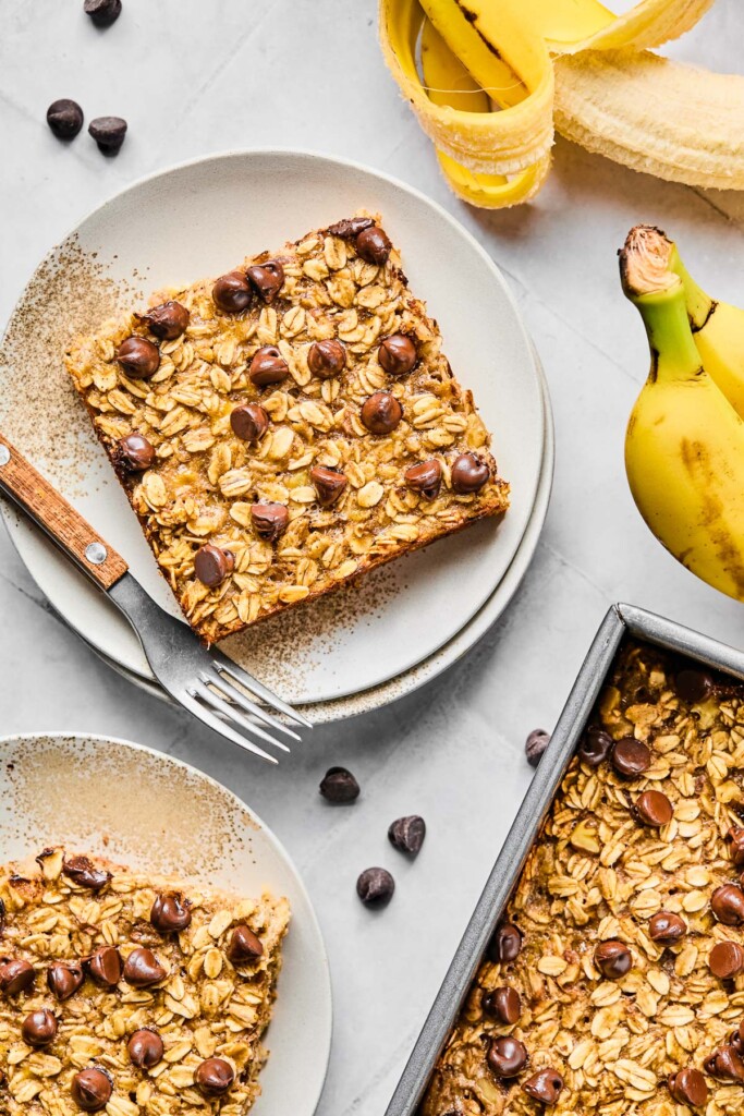 Overhead shot of two pieces of banana bread baked oatmeal on white plates and a pan of baked oatmeal next to them. One of the plates has a fork on it. Around the dishes are some chocolate chips and bananas.