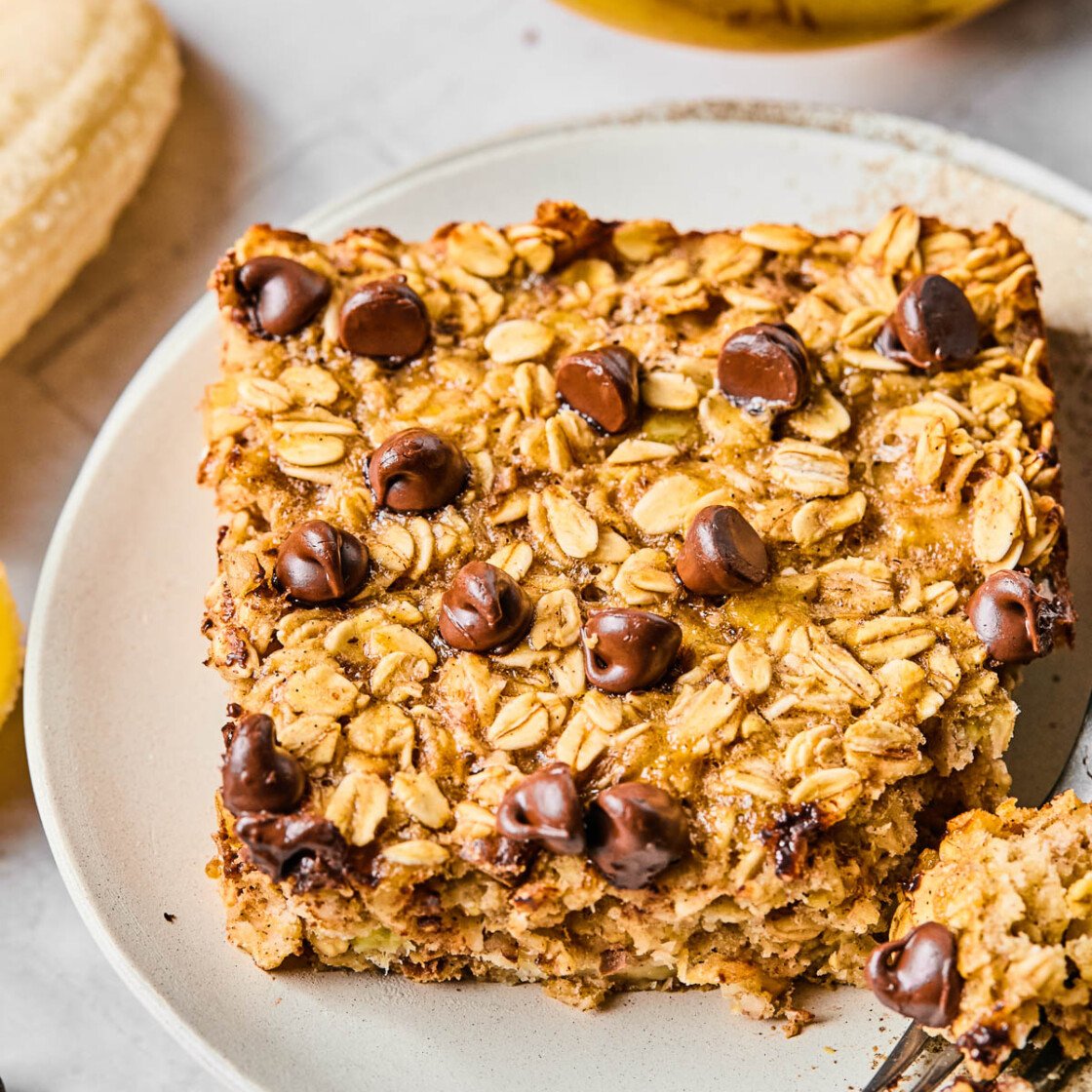 A piece of banana baked oatmeal with chocolate chips on a white plate. A little piece is taken off the corner and sitting on a fork on the plate. Around the plate are some chocolate chips and some bananas.