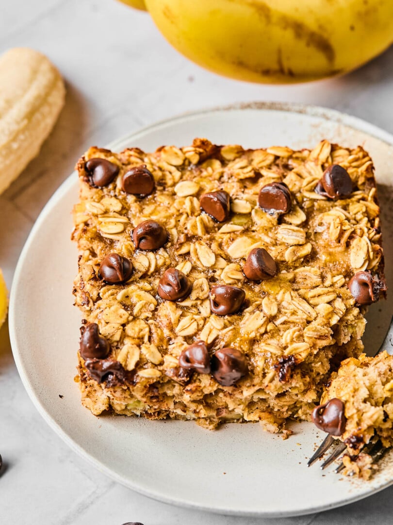 A piece of banana baked oatmeal with chocolate chips on a white plate. A little piece is taken off the corner and sitting on a fork on the plate. Around the plate are some chocolate chips and some bananas.