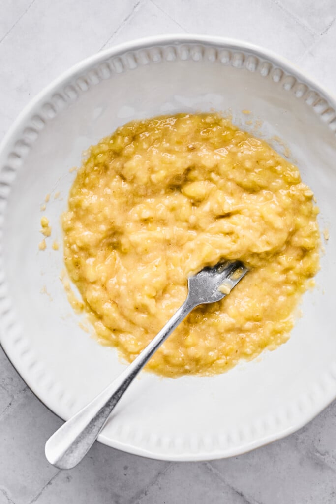 A white bowl of mashed bananas with a fork in it.
