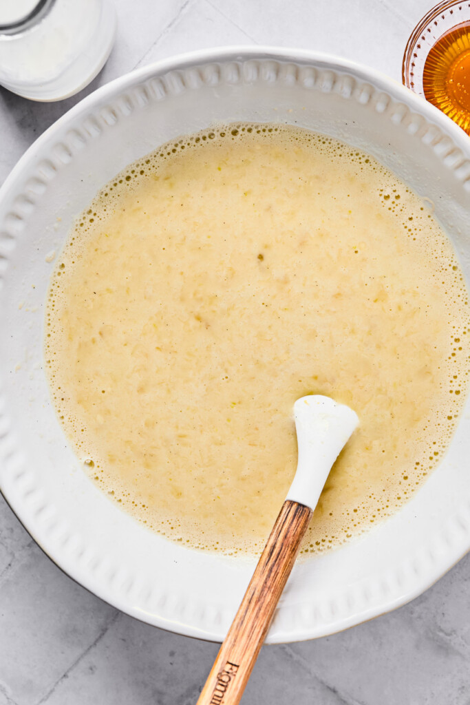 Large white mixing bowl with a wet mixture of eggs, milk, mashed bananas, honey, and vanilla in it. There is a spatula in the bowl as well. Around the bowl is a jar of milk and a small bowl of honey.
