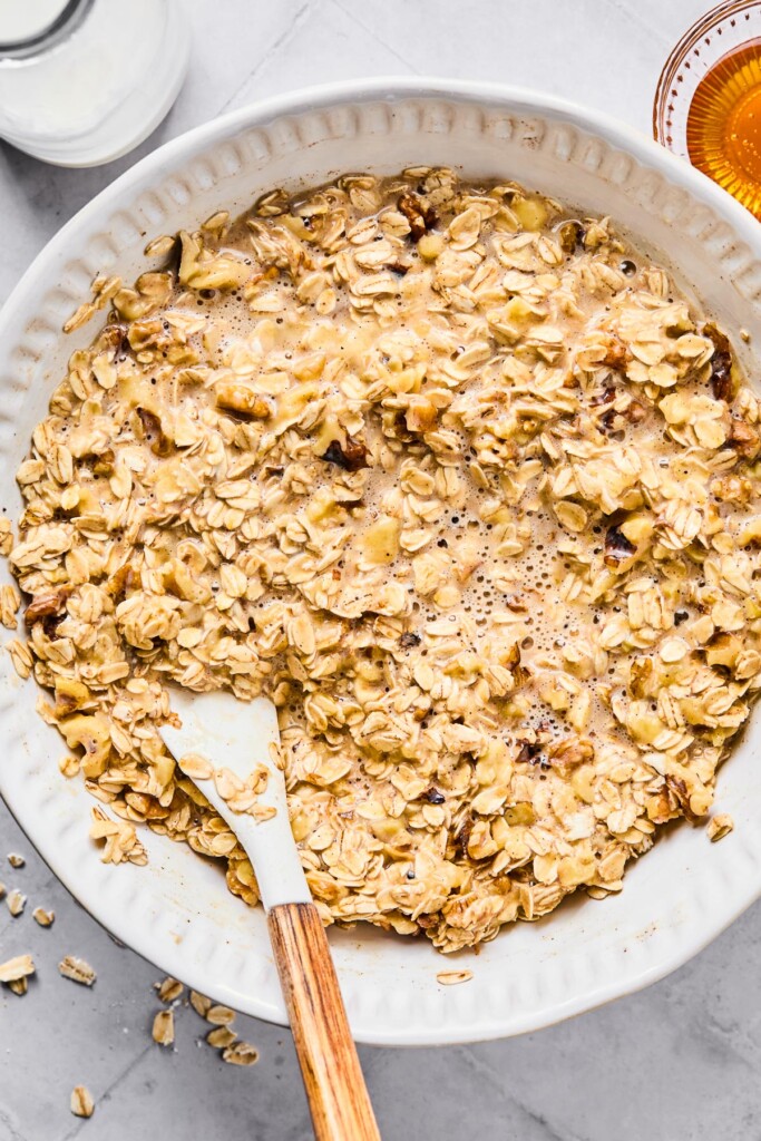A white mixing bowl filled with a wet batter of milk, eggs, rolled oats, walnuts, and spices. A spatula is also in the bowl. Around the bowl is a jar of milk and a small bowl of honey.