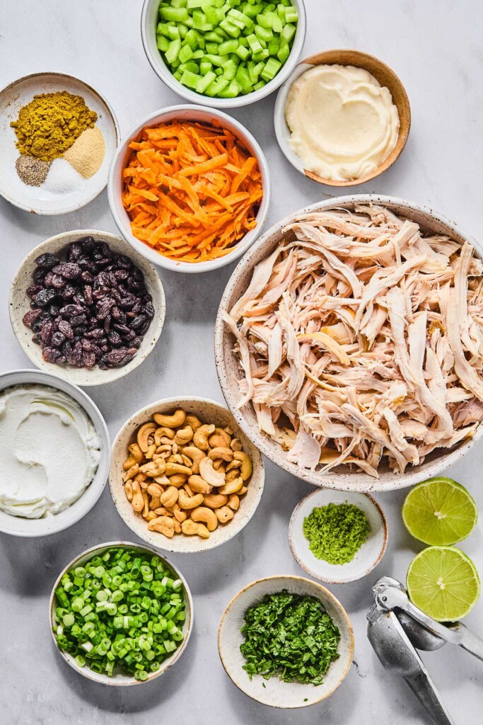 A bowl of diced celery, a bowl of mayo, a large bowl of shredded chicken, a lime that has been cut in half, a metal citrus press, a small bow of lime zest, a bowl of chopped cilantro, a bowl of chopped green onions, a bowl of cashews, a bowl of greek yogurt, a bowl of raisins, a bowl of shredded carrots, and a bowl of a few spices in separate piles on a marble counter.