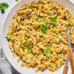A large bowl of curry chicken salad with raisins, cashews, and veggies, garnished with cilantro. There is a spoon and a fork in the bowl as well. Around the bowl is a striped kitchen towel, some cilantro leaves, and some slices of bread.