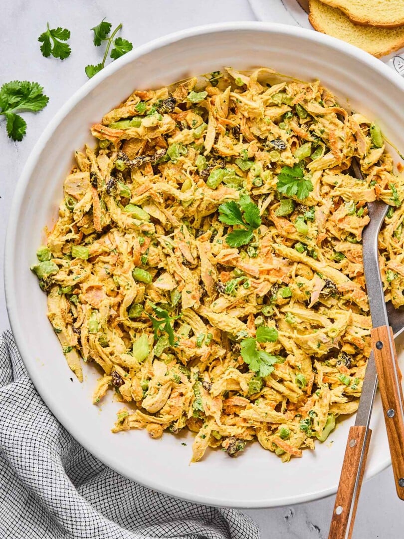 A large bowl of curry chicken salad with raisins, cashews, and veggies, garnished with cilantro. There is a spoon and a fork in the bowl as well. Around the bowl is a striped kitchen towel, some cilantro leaves, and some slices of bread.