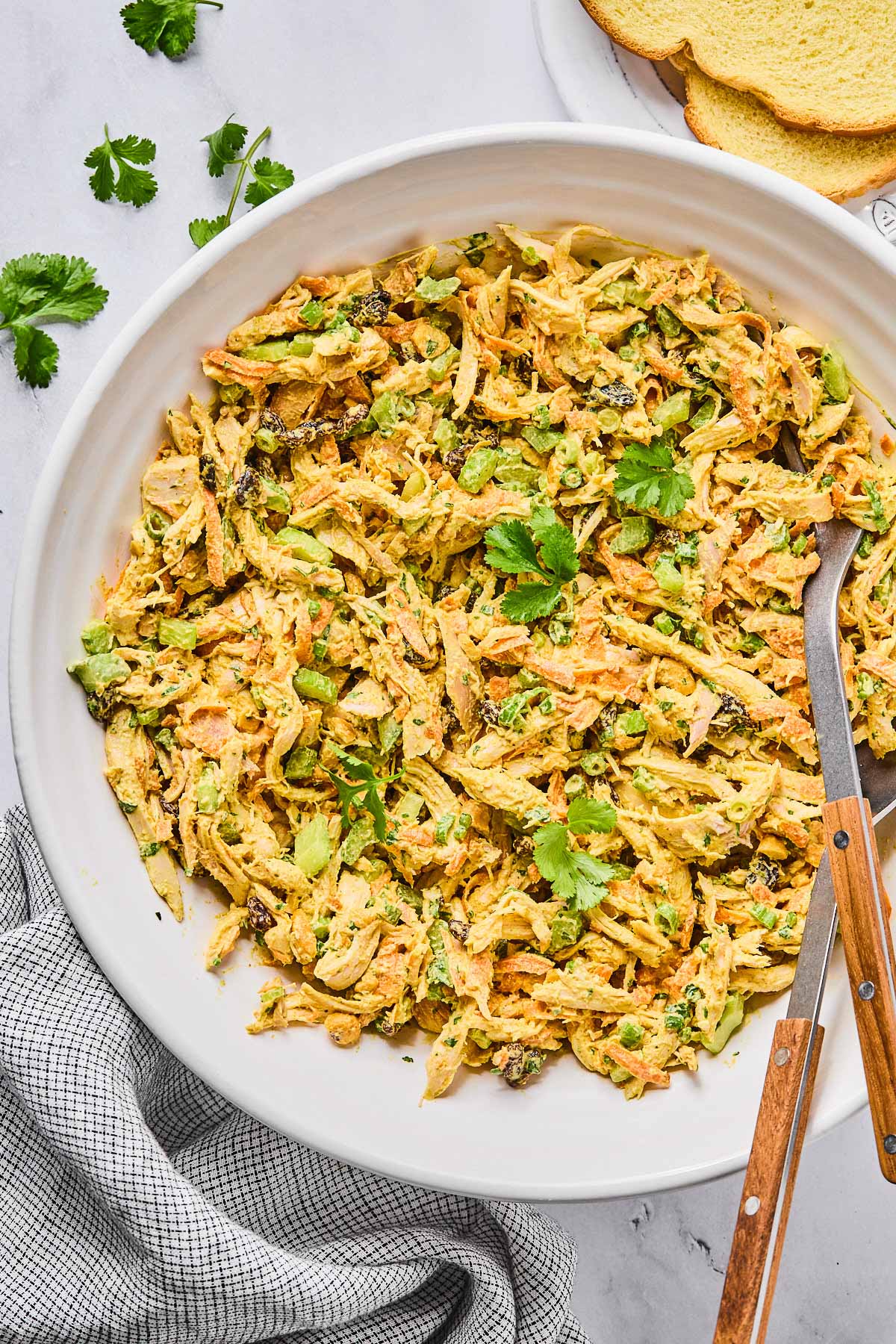 A large bowl of curry chicken salad with raisins, cashews, and veggies, garnished with cilantro. There is a spoon and a fork in the bowl as well. Around the bowl is a striped kitchen towel, some cilantro leaves, and some slices of bread.