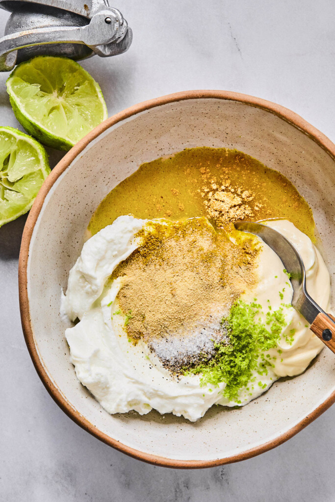 Overhead shot of a bowl with greek yogurt, mayo, lime juice, lime zest, spices, salt, and pepper in it before it is mixed together. A spoon is in the bowl as well. Next to the bowl is a lime that has been cut in half and juiced and a metal citrus press.