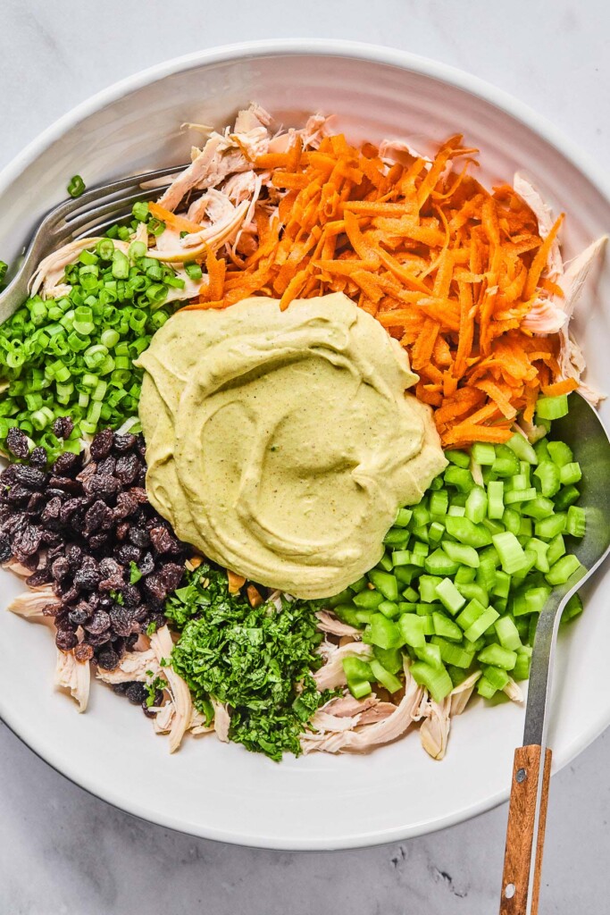 A large white bowl with shredded chicken topped with a pile of shredded carrots, a pile of diced celery, a pile of chopped cilantro, a pile of raisins, a pile of chopped green onions, and a spiced yogurt sauce before it is mixed together. A spoon is in the bowl as well.