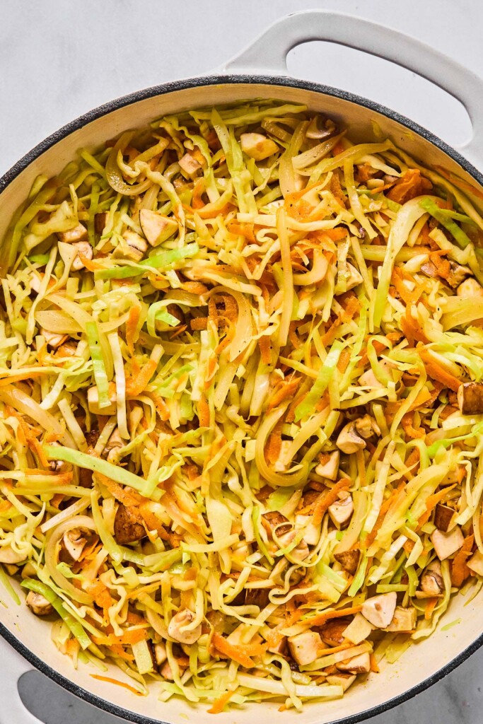 Overhead shot of a large white enameled pan filled with sautéed sliced cabbage, shredded carrots, and diced mushrooms.