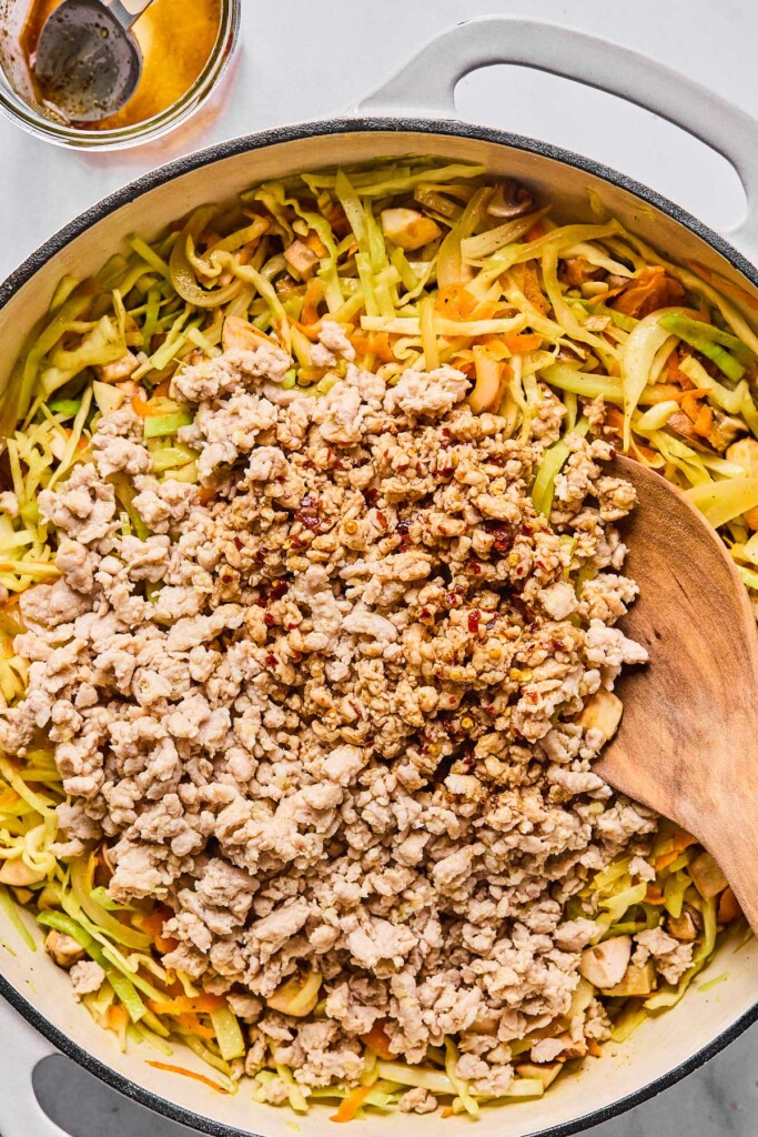 Overhead shot of a large white enameled pan filled with sautéed ground chicken, sliced cabbage, shredded carrots, and diced mushrooms with a pile of cooked ground chicken and stir fry sauce on top of it. A wooden spoon is in the pan as well. Next to the pan is a small jar with a spoon in it that the stir fry sauce was in.