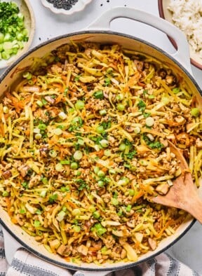 Healthy egg roll in a bowl in a large white skillet. It is garnished with black sesame seeds, sliced green onions, and cilantro and a wooden spoon is in the pan as well. Around the pan is a white and blue kitchen towel, a bowl with cilantro and sliced green onions, a small bowl of black sesame seeds, and a bowl of white rice.