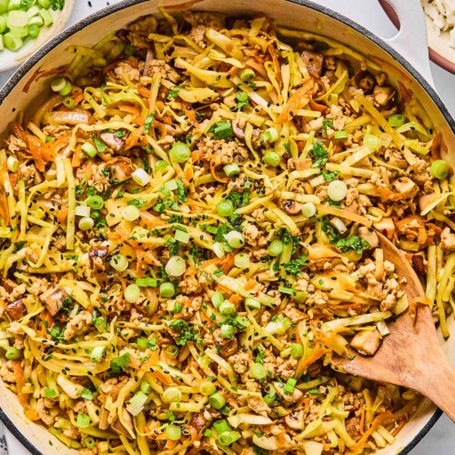 Healthy egg roll in a bowl in a large white skillet. It is garnished with black sesame seeds, sliced green onions, and cilantro and a wooden spoon is in the pan as well. Around the pan is a white and blue kitchen towel, a bowl with cilantro and sliced green onions, a small bowl of black sesame seeds, and a bowl of white rice.