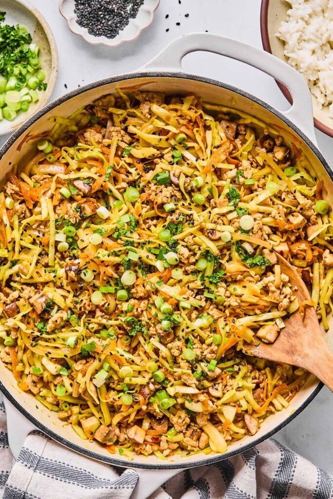 Healthy egg roll in a bowl in a large white skillet. It is garnished with black sesame seeds, sliced green onions, and cilantro and a wooden spoon is in the pan as well. Around the pan is a white and blue kitchen towel, a bowl with cilantro and sliced green onions, a small bowl of black sesame seeds, and a bowl of white rice.