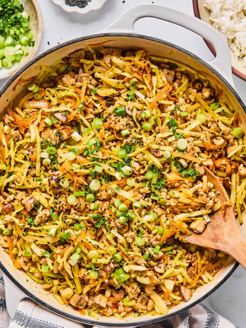 Healthy egg roll in a bowl in a large white skillet. It is garnished with black sesame seeds, sliced green onions, and cilantro and a wooden spoon is in the pan as well. Around the pan is a white and blue kitchen towel, a bowl with cilantro and sliced green onions, a small bowl of black sesame seeds, and a bowl of white rice.