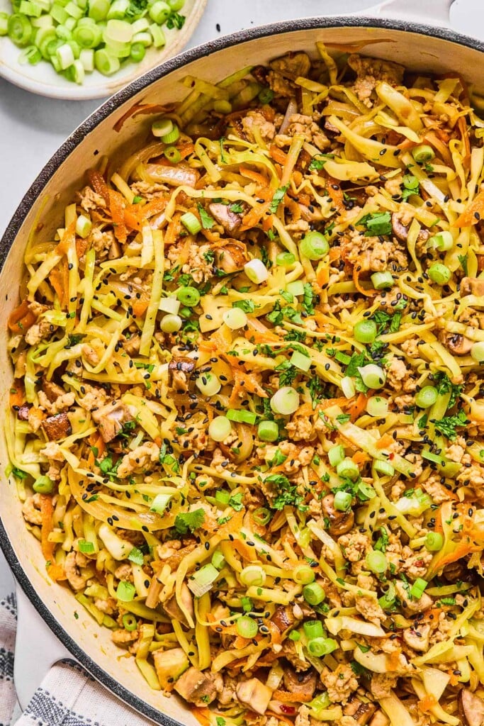 A close up of a pan of egg roll in a bowl topped with sliced green onions, cilantro, and black sesame seeds. Next to the pan is a bowl of sliced green onions.