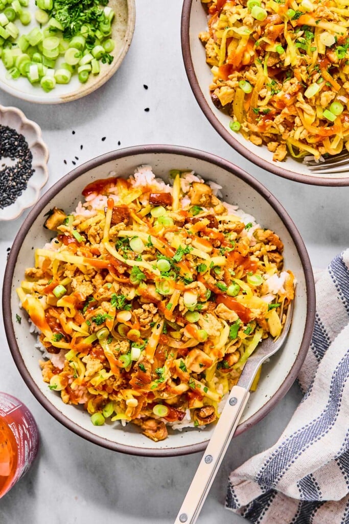 Bowl bowls of chicken egg roll in a bowl over white rice. They are topped with sriracha, sliced green onions, cilantro, and sesame seeds. A fork is in one of the bowls. Around the bowls is a white and blue kitchen towel, a container of sriracha, a bowl of black sesame seeds, and a bowl of sliced green onions and cilantro.