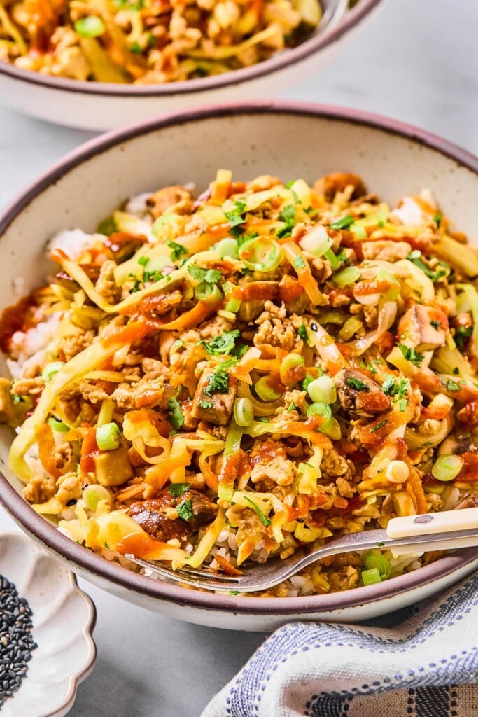 Egg roll bowl with ground chicken, sliced cabbage, shredded carrots, and mushrooms over white rice. It is in a bowl and garnished with sriracha, green onions, cilantro, and sesame seeds. A fork is also in the bowl. Around the bowl is a kitchen towel, a bowl of black sesame seeds, and another egg roll bowl.