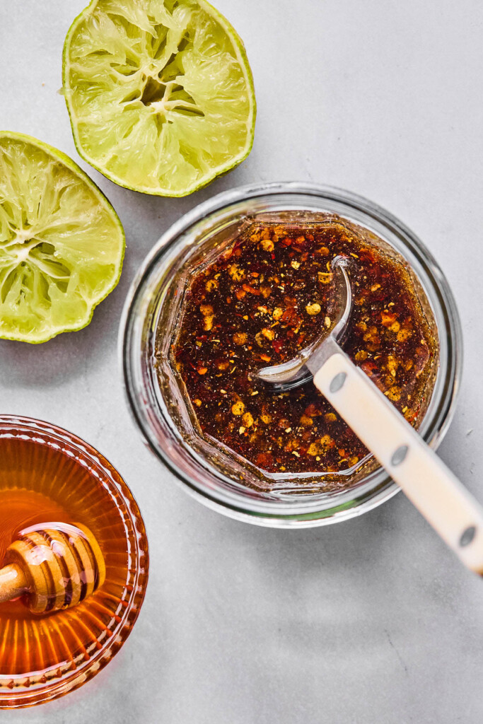 Small jar of stir fry sauce with a spoon in it. Next to it is a lime that has been cut in half and juiced and a small bowl of honey with a honey dipper in it.