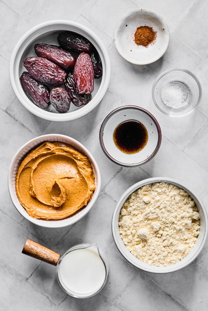 A small bowl of cinnamon, a small bowl of salt, a small bowl of vanilla bean paste, a bowl of vanilla protein powder, a measuring cup of milk, a bowl of peanut butter, and a bowl of medjool dates on a grey tile counter.