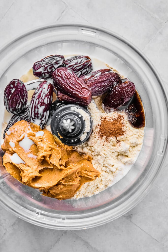 The bowl of a food processor filled with medjool dates, vanilla bean paste, protein powder, cinnamon, peanut butter, and milk before it is processed together.