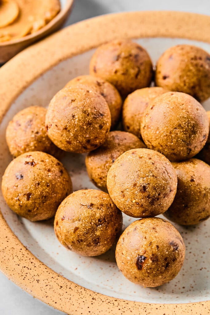 A close up of protein peanut butter balls stacked on top of each other on a white and brown plate. Behind the plate is a bowl of peanut butter.
